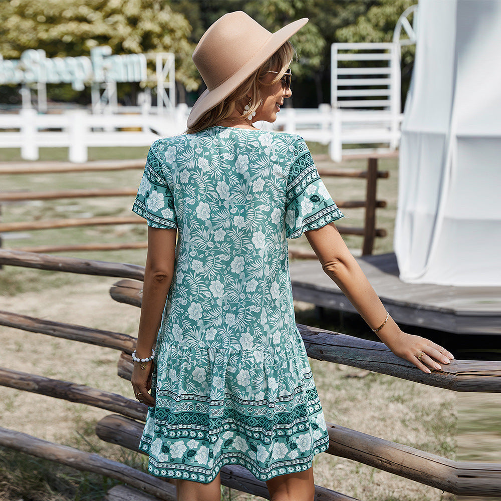 Woman in a green floral dress and beige hat standing by a wooden fence outdoors.
